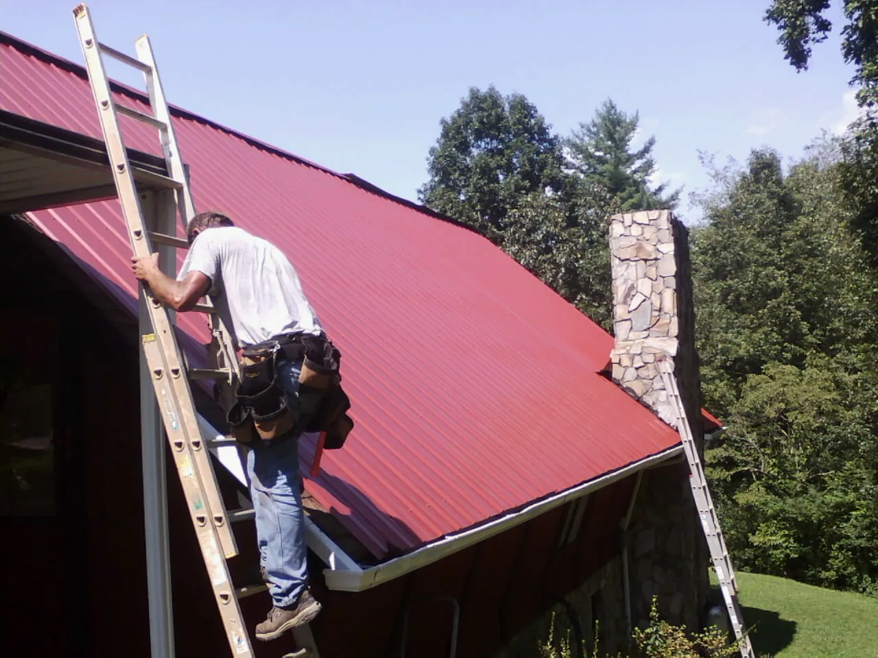 Roofer on a ladder beside a red metal roof and stone chimney — Soffit & Fascia Repair by Roofing Brothers in Anoka